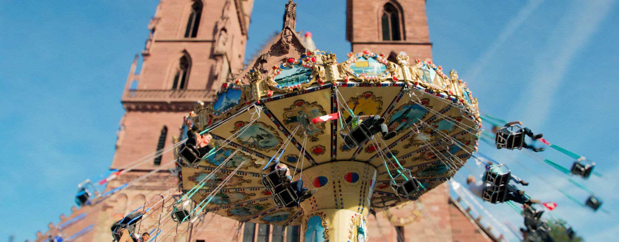 A cheerful carousel with colourful horses and other rides turns in front of the imposing Elisabethenkirche in Basel. Children and adults laugh and wave as they make their rounds. The church rises majestically in the background, with its tall bell tower and richly decorated façade.